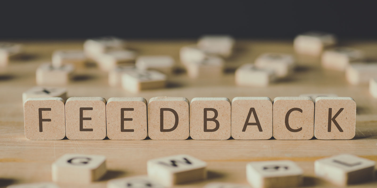 Wooden letter blocks spelling “FEEDBACK” arranged on a table, with scattered blocks blurred in the background.