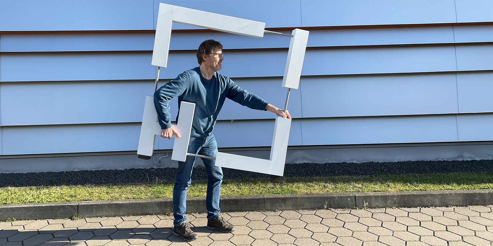Person standing on a paved walkway outside a modern building, holding a large lightweight square frame made of white rectangular elements and the letters "H" and "I" attached to metal rods.