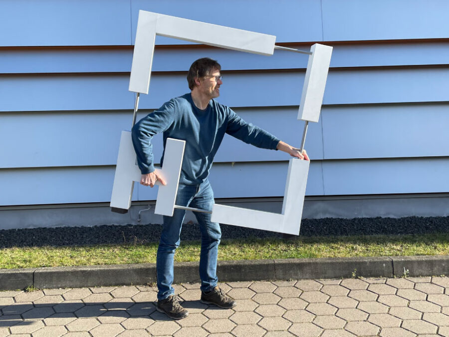 Person standing on a paved walkway outside a modern building, holding a large lightweight square frame made of white rectangular elements and the letters "H" and "I" attached to metal rods.
