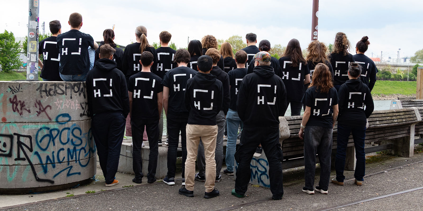 Group of Helmholtz Imaging team standing outdoors with their backs to the camera, all wearing matching black T-shirts and hoodies with the Helmholtz Imaging logo.