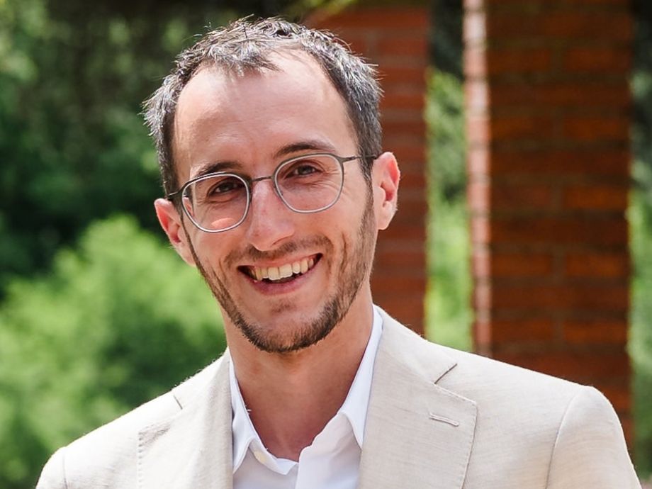 Portrait of Manuel Haussmann smiling at the camera, wearing glasses, a light beige blazer and a white shirt, with greenery and brick pillars in the background.